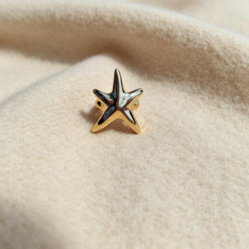 A gold starfish-shaped ring with a polished silver-toned top, resting on a soft, textured beige fabric background.