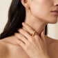 A close-up lifestyle shot of a model wearing a gold starfish ring on her finger and a matching gold bar earring in her ear. The image features warm, soft lighting against a neutral beige background.