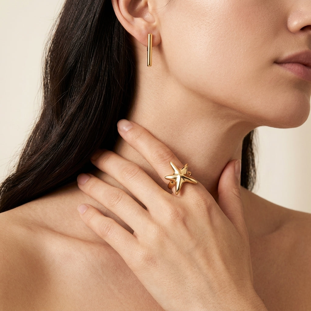 A close-up lifestyle shot of a model wearing a gold starfish ring on her finger and a matching gold bar earring in her ear. The image features warm, soft lighting against a neutral beige background.