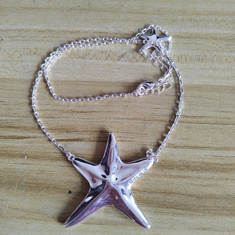 A silver starfish pendant necklace with a matching starfish charm on a silver chain, displayed on a light wood grain surface.