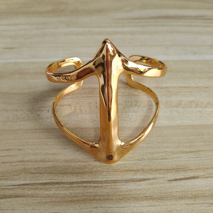 A polished gold-tone metal ring with a sculptural, openwork design resembling a stylized anchor or cross, displayed on a light wood grain surface.