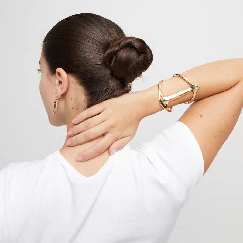A woman with her hair in a bun is shown from the back wearing a chunky, geometric gold cuff bracelet and a small gold hoop earring, holding her neck against a light gray background.