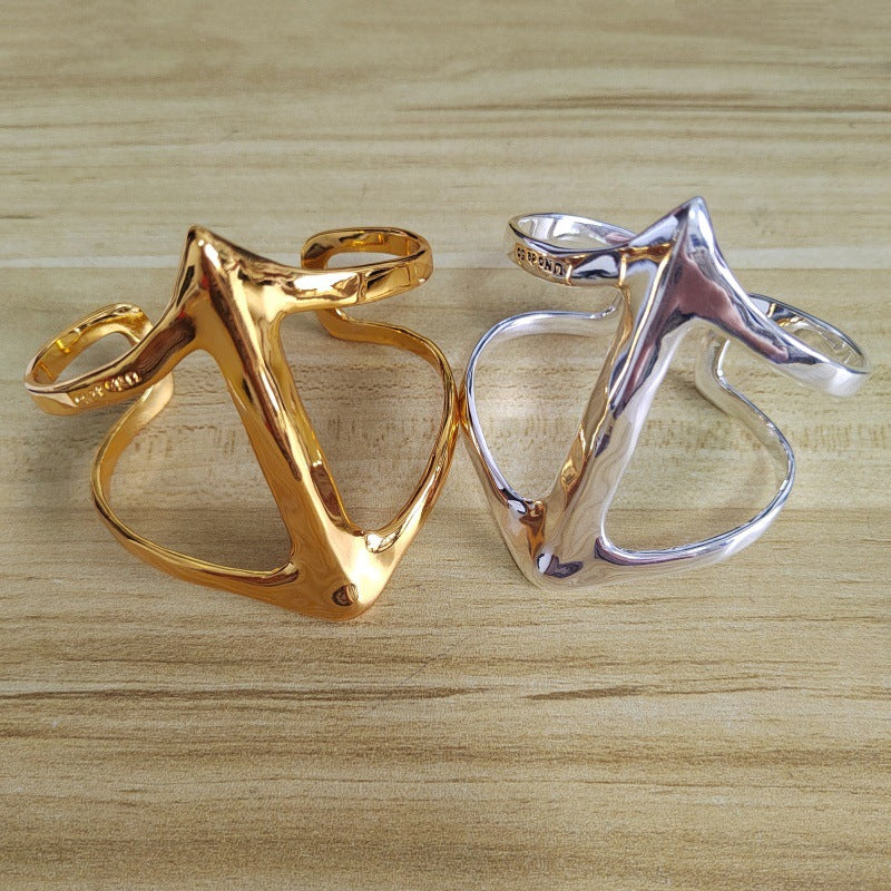 Two abstract, organic-shaped ring bangles displayed side-by-side on a light wood grain surface. One is polished gold and the other is polished silver.