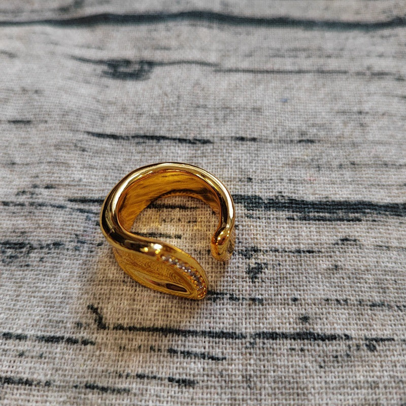 A gold-colored open cuff ring with a textured, hammered finish and a small row of clear stones on one side, resting on a gray textured fabric background.