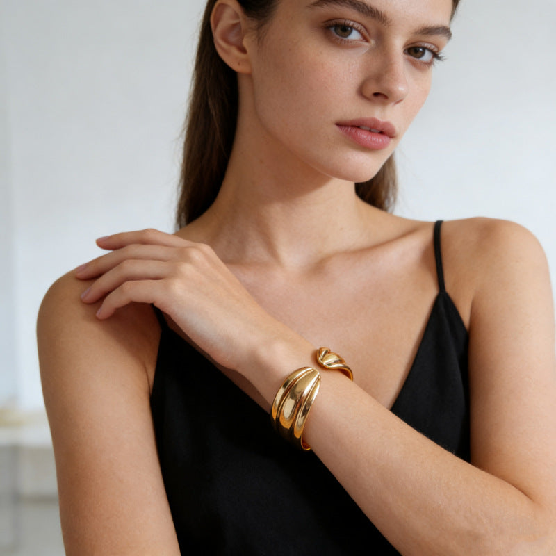 A close-up of a woman wearing a polished gold wrap bracelet on her wrist, dressed in a black spaghetti-strap top against a soft, light gray background.