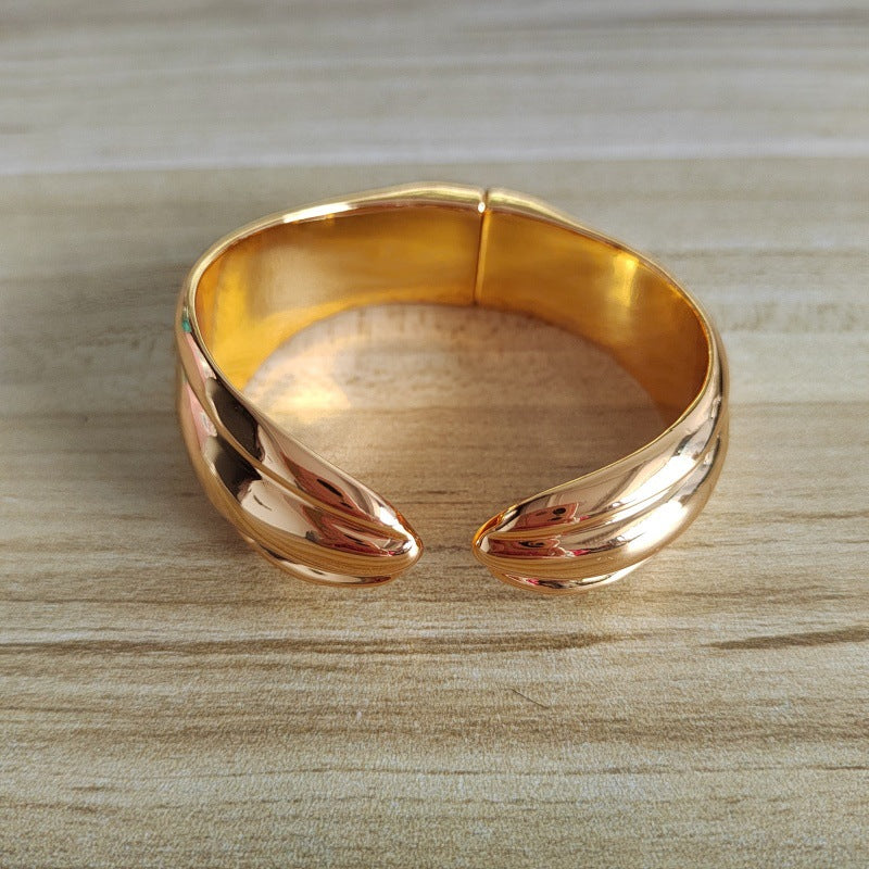 A polished rose gold cuff bracelet with a twisted, leaf-like design, resting on a light wood grain surface.