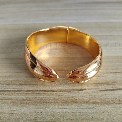 A polished rose gold cuff bracelet with a twisted, leaf-like design, resting on a light wood grain surface.