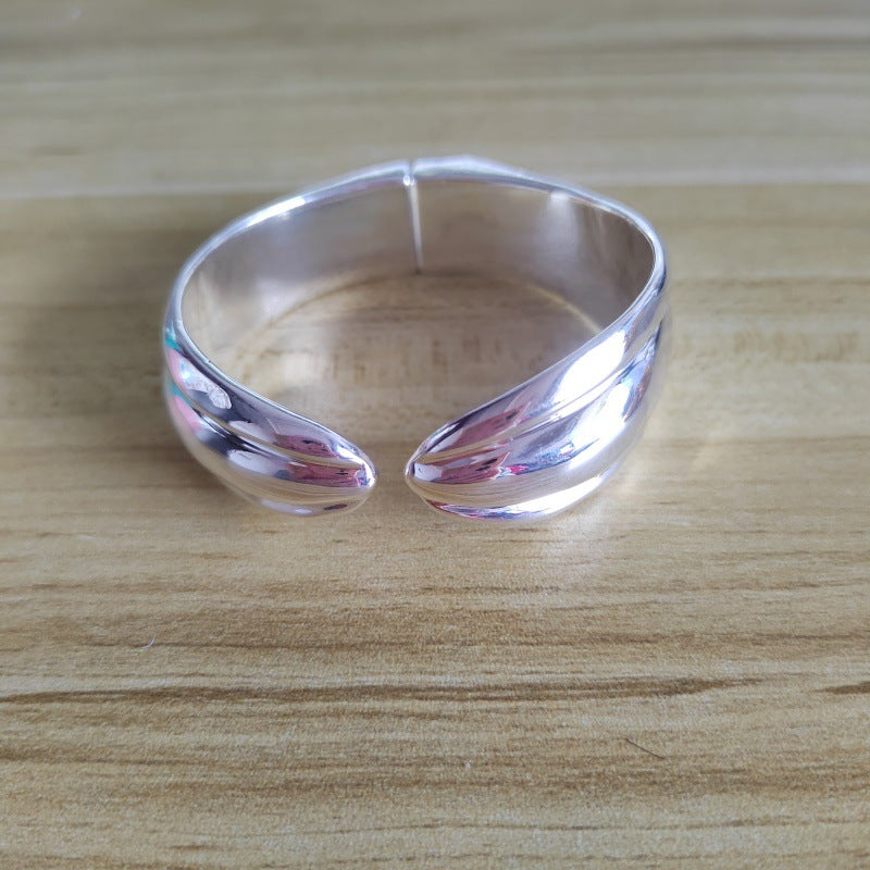 A polished silver open cuff ring with a smooth, curved, wave-like design, resting on a light brown wooden surface.