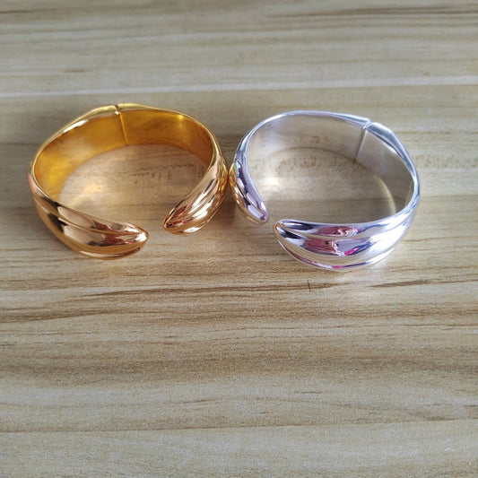 Two metallic open cuff rings, one in polished gold and one in polished silver, displayed side-by-side on a light wood grain surface.