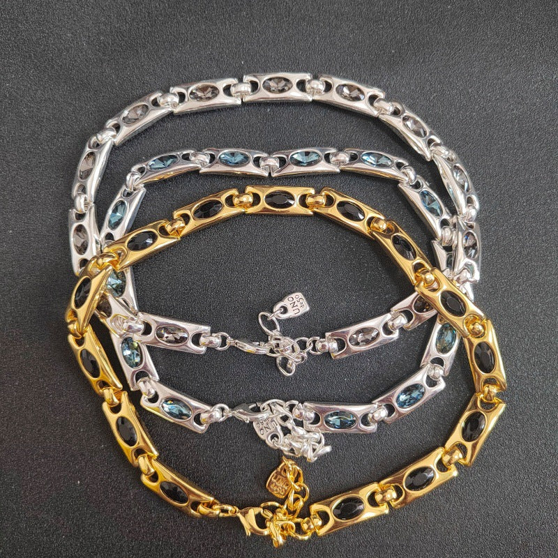 Four chain bracelets stacked on a dark gray background, featuring alternating links of silver, yellow gold, and dark gunmetal gray, with some links inlaid with oval blue-green stones.