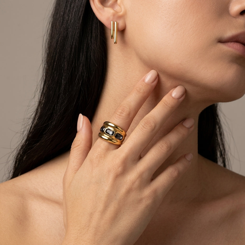 A close-up lifestyle shot of a model wearing a gold ring with black enamel details on her finger, along with a matching gold hoop earring, set against a neutral beige background.