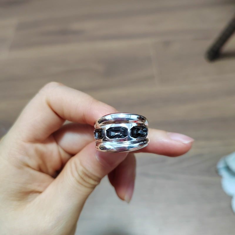 A hand holding a wide silver ring with three dark oval gemstones, set against a blurred wooden floor background.