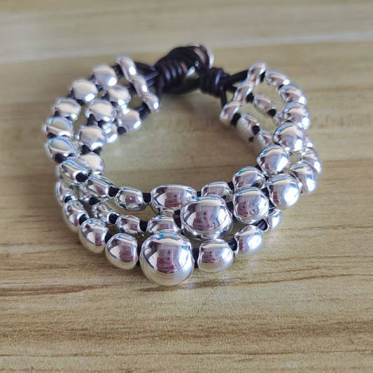 A multi-strand silver beaded bracelet with a dark brown cord closure, displayed on a light wood grain surface.