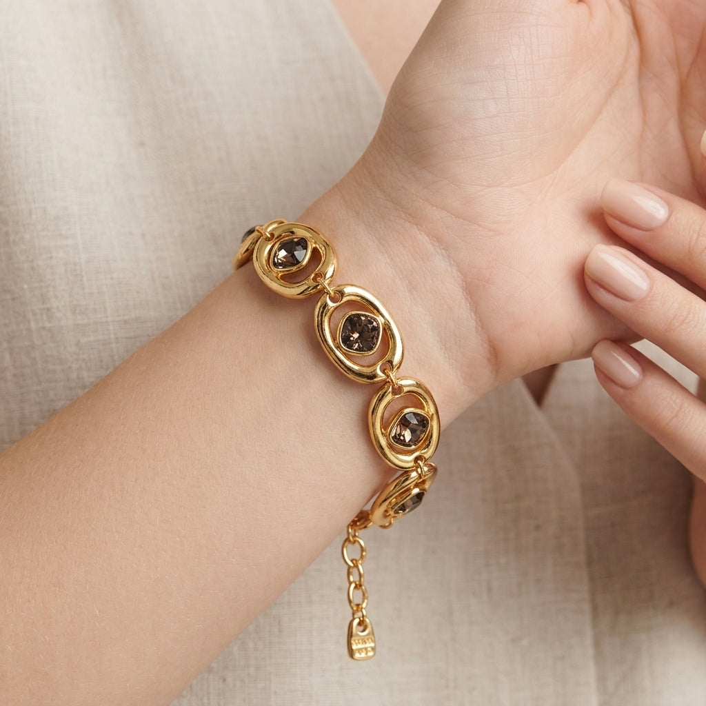 A gold-toned bracelet with three oval links, each featuring a dark brown smoky gemstone, shown worn on a woman's wrist against a neutral beige linen background.