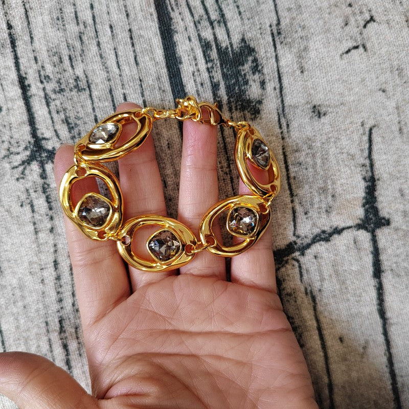 A gold chain bracelet with oval links and smoky brown gemstone accents, shown resting on a person's hand against a textured gray fabric background.
