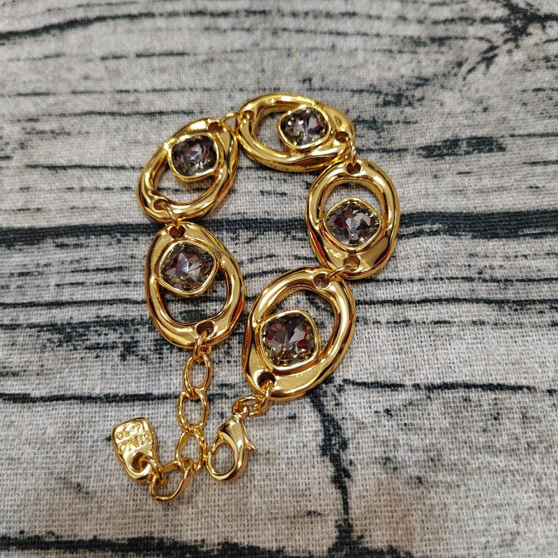 A gold-tone bracelet with five oval links, each featuring a faceted smoky purple gemstone, displayed on a textured gray fabric background.