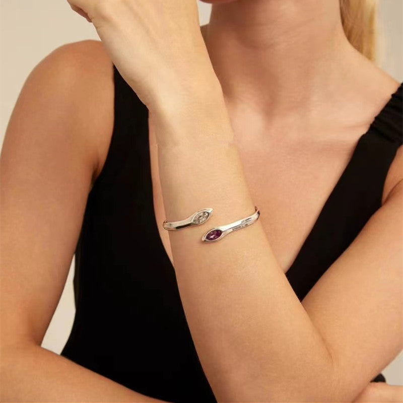 A close-up of a woman's forearm wearing a silver-toned open cuff bracelet with one end featuring a small clear stone and the other a teardrop-shaped purple gemstone, paired with a black sleeveless top.