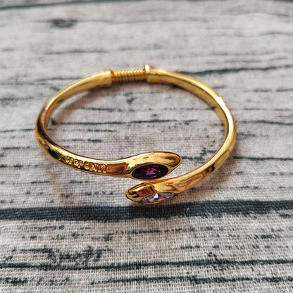 A gold-plated snake-shaped cuff bracelet with two oval gemstone accents, one dark purple and one clear, resting on a gray textured fabric background.