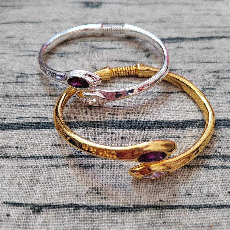 Two open cuff bracelets, one silver-toned and one gold-toned, each featuring a polished snake head motif and a small oval purple gemstone, displayed on a textured gray fabric background.