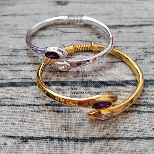 Two open cuff bracelets, one silver-toned and one gold-toned, each featuring a polished snake head motif and a small oval purple gemstone, displayed on a textured gray fabric background.