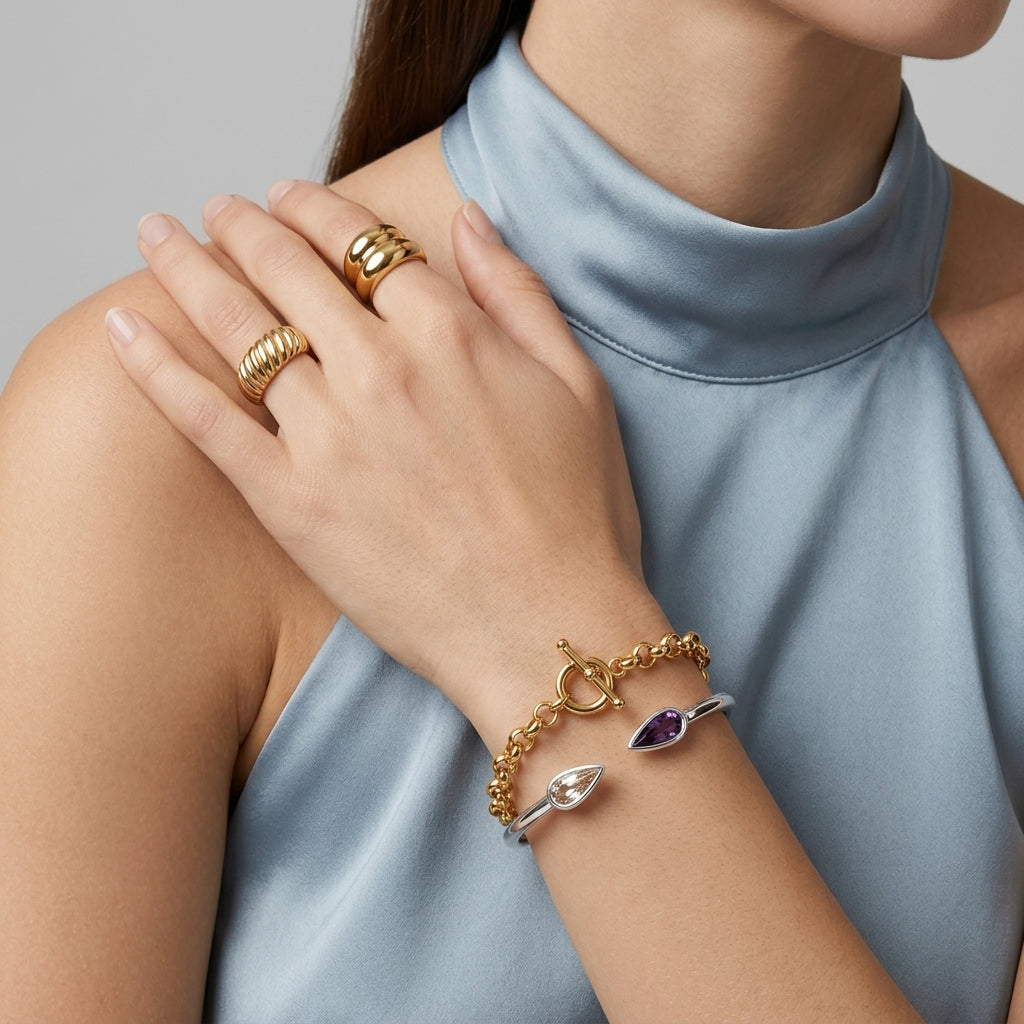 A close-up of a model wearing multiple gold jewelry pieces, including stacked rings on one finger and a gold chain bracelet with a toggle clasp and two accent charms on the wrist. The model is wearing a light blue satin high-neck top against a neutral gray background.