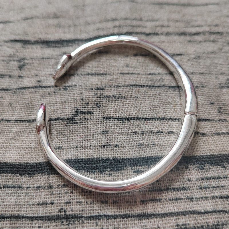 A polished silver-toned metal hinged cuff bracelet with a split clasp design, resting on a textured gray and black striped fabric background.