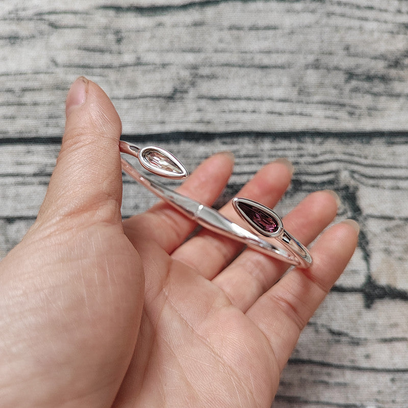 A hand holding a silver-toned open cuff bracelet with two marquise-cut gemstones, one clear and one dark pinkish-red, against a gray wood-textured background.