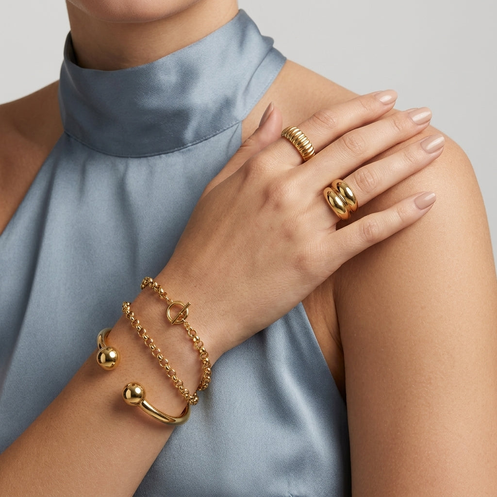 A close-up of a model's hand and wrist wearing multiple gold jewelry pieces, including two rings and three bracelets, styled with a high-neck satin blue top against a neutral gray background.