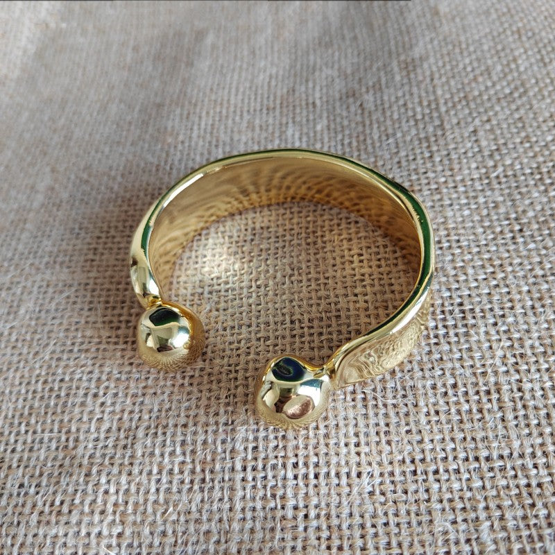 A gold-toned open cuff bracelet with two small dark oval inlays, photographed on a textured beige fabric background.