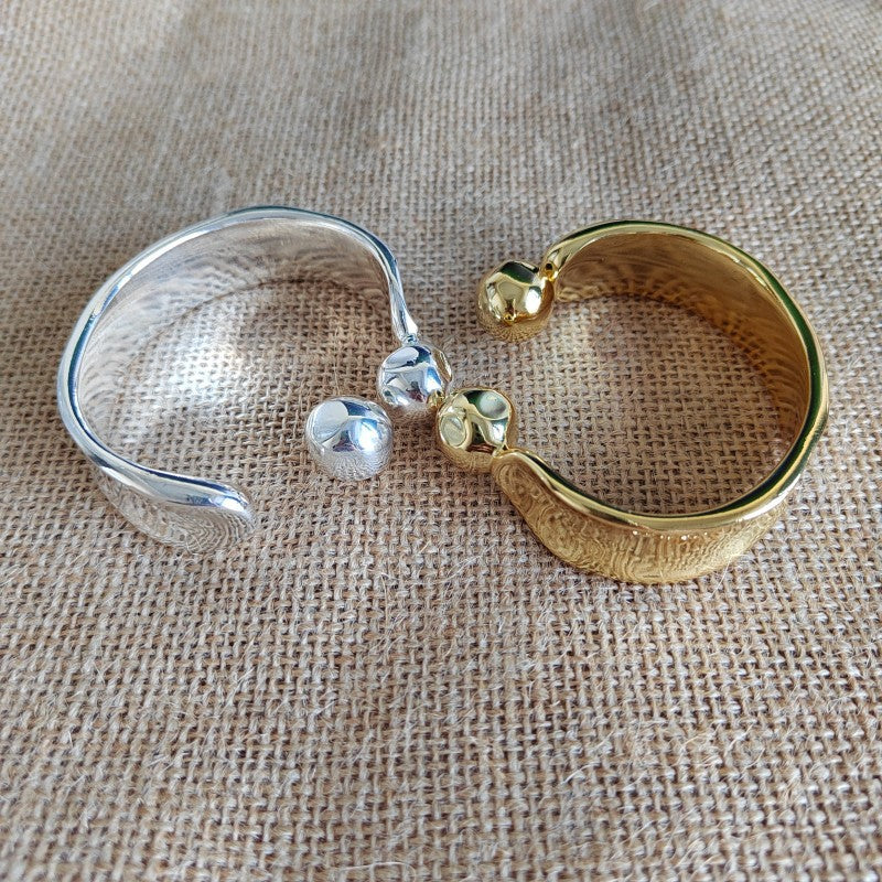 Two textured open cuff bracelets, one silver-toned and one gold-toned, each with a small bead detail, displayed on a beige burlap fabric background.