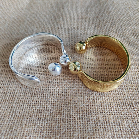 Two textured open cuff bracelets, one silver-toned and one gold-toned, each with a small bead detail, displayed on a beige burlap fabric background.