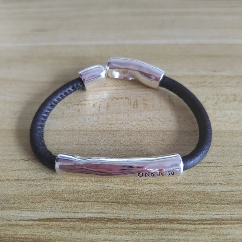 A silver-toned UN920 bracelet with a textured horizontal bar and polished end caps, paired with a black leather cord, displayed on a light wood grain surface.