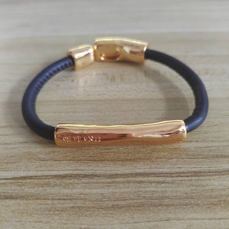 A dark navy blue cord bracelet with polished rose gold metal accents, including a central bar and clasp, displayed on a light wood grain surface.