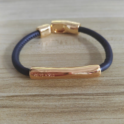 A dark navy blue cord bracelet with polished rose gold metal accents, including a central bar and clasp, displayed on a light wood grain surface.