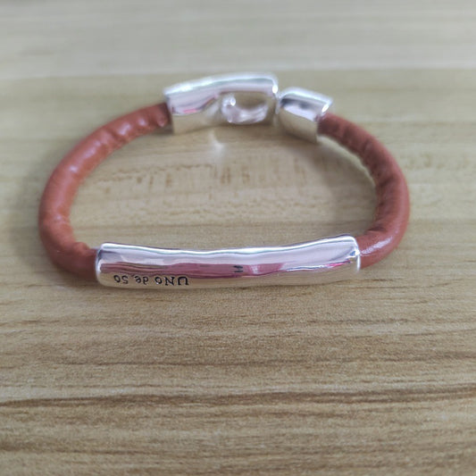A silver-toned bar bracelet with a reddish-brown leather wrap, resting on a light wood grain surface.