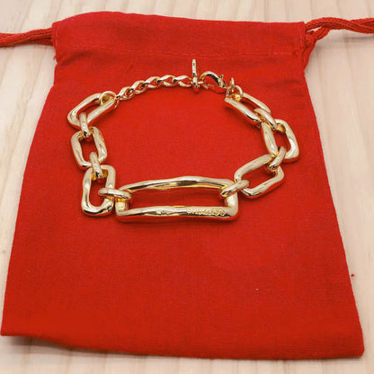 A chunky gold chain bracelet with a large horizontal bar link, displayed on a red drawstring pouch over a light wood surface.