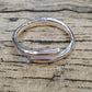 A polished sterling silver open bangle bracelet with a smooth, rounded design, resting on a textured beige linen fabric with black wood grain patterns.