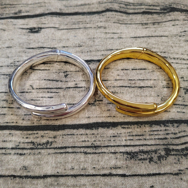 Two polished metal bangle bracelets, one silver-toned and one gold-toned, displayed side-by-side on a textured beige fabric with a dark wood grain pattern.
