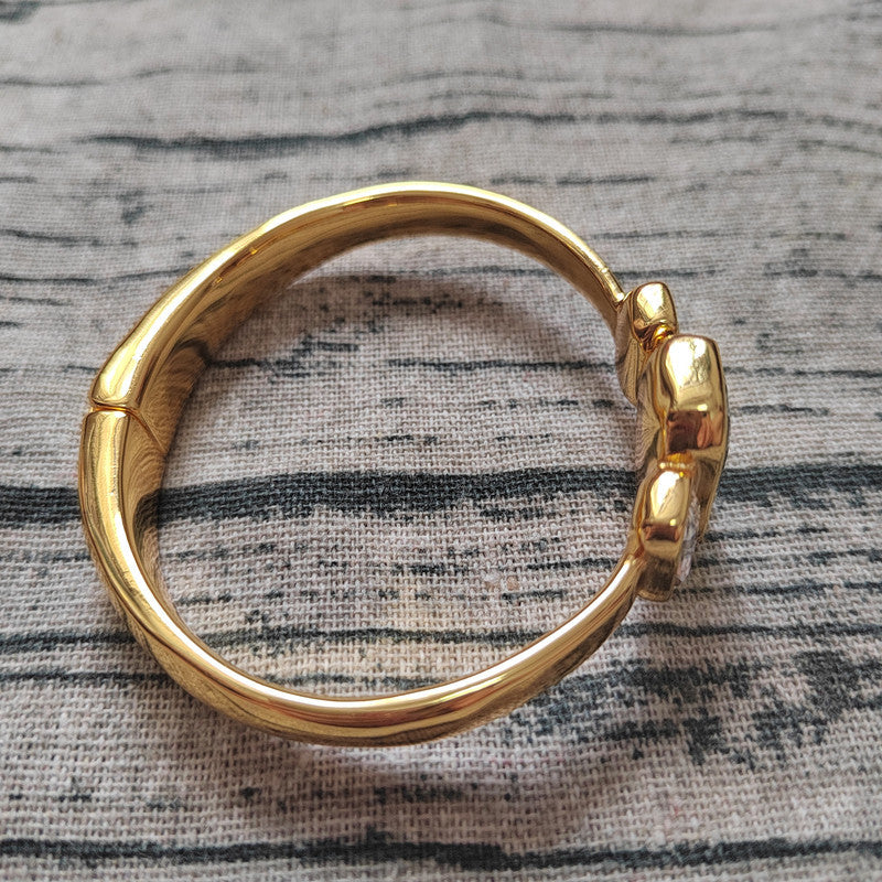 A polished gold-colored metal hoop earring with a textured, organic curved design and a small raised decorative element on the front, displayed on a gray and black patterned fabric background.