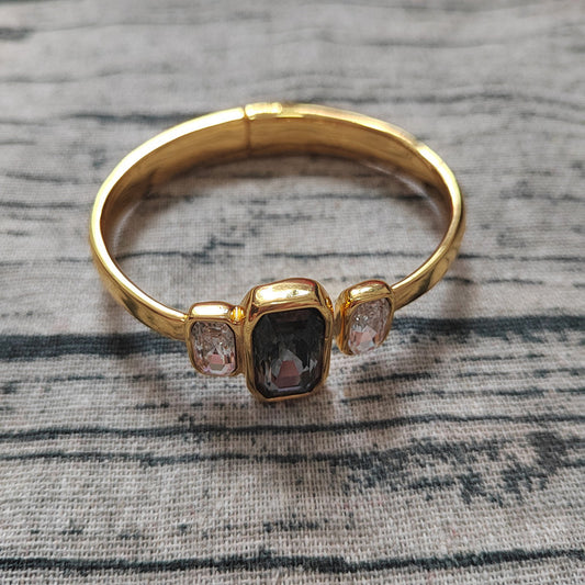 A gold bangle ring with a central rectangular dark gray stone and two small clear stones on either side, resting on a textured gray fabric background.