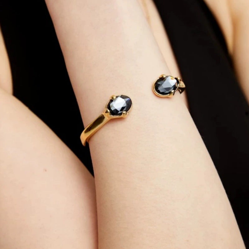 A close-up of a person's wrist wearing a gold ring with a faceted dark gray gemstone, against a black top background.
