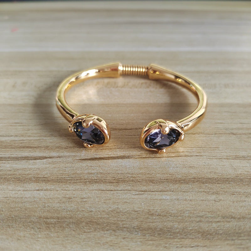 A gold-plated open cuff bracelet with two oval bezel-set dark purple gemstones, displayed on a light wood grain surface.