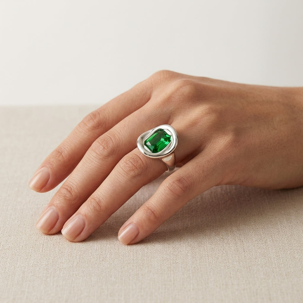 A silver ring with a cushion-cut green emerald gemstone, shown worn on a hand resting on a neutral beige textured surface.