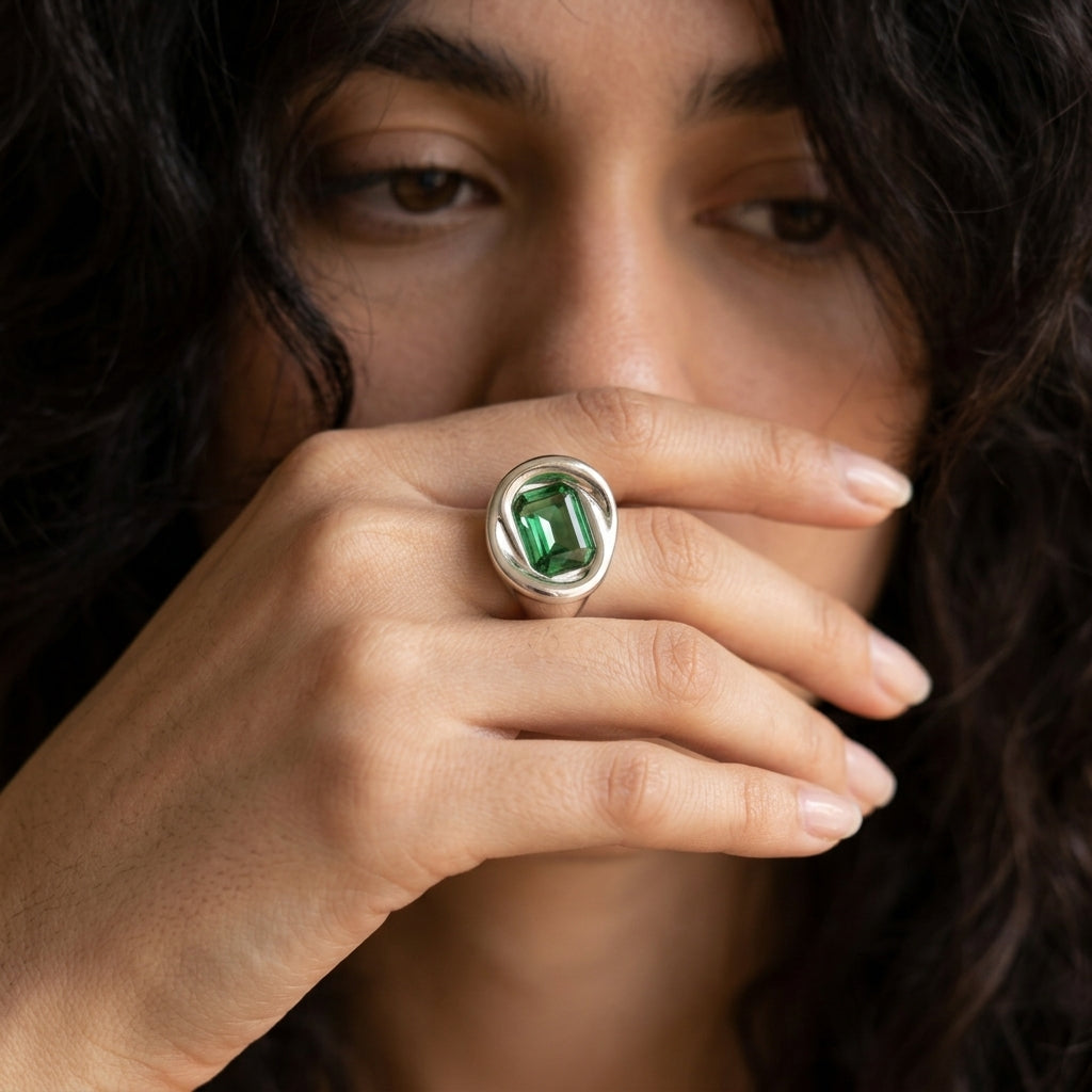 A close-up lifestyle shot of a woman wearing a silver ring with a green emerald-cut gemstone, holding a cup near her hand.