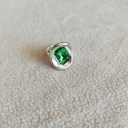 A silver ring featuring a large, cushion-cut green gemstone, resting on a textured beige fabric surface.