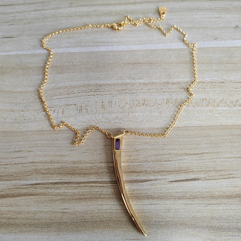 A gold chain necklace with a curved, horn-like pendant featuring a small purple rectangular accent, displayed on a light wood grain surface.