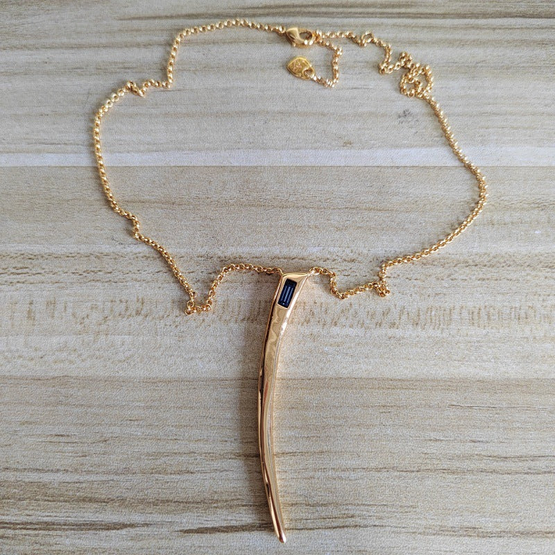 A gold chain necklace with a curved, polished gold pendant featuring a small rectangular dark blue inlay, displayed on a light wood grain surface.