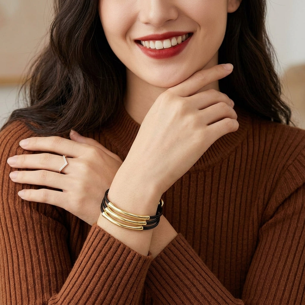A close-up lifestyle shot of a woman wearing a stack of three gold bangle bracelets with a black wrap, paired with a brown ribbed sweater.