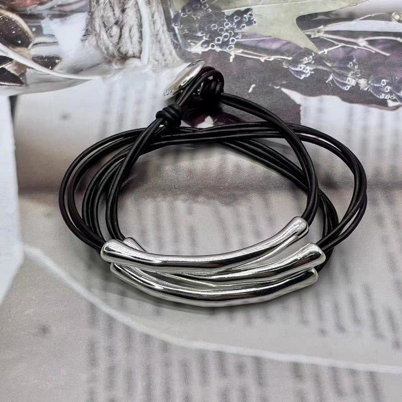 A multi-strand black cord bracelet with three polished silver-toned curved bar charms, displayed on a light gray textured surface.