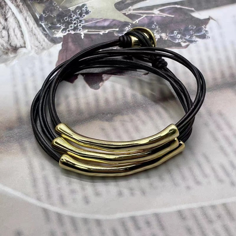 A multi-strand black cord bracelet with three polished gold-tone curved bar accents at the front, displayed on a vintage-style printed paper background.